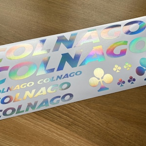 Puede incluir: Una hoja de pegatinas de vinilo blanco con letras holográficas iridiscentes que dicen "COLNAGO" en varios tamaños y estilos. También hay cuatro pequeños diseños de trébol de cuatro hojas holográficos iridiscentes.