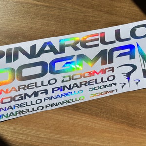 以下が含まれることがあります： 虹色のホログラフィックフォントで「Pinarello Dogma」という単語が複数回繰り返された白いステッカーです。