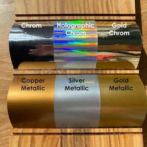 May include: Six samples of vinyl adhesive film in different colors and finishes. From left to right: Chrome, Holographic Chrome, Gold Chrome, Copper Metallic, Silver Metallic, and Gold Metallic.