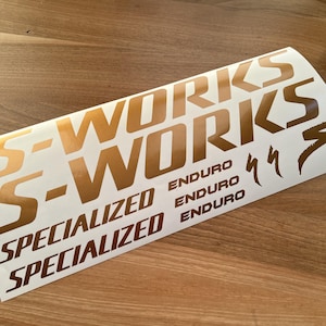 May include: A sheet of gold-coloured Specialized S-Works Enduro decals. The decals are arranged in multiple rows, with the brand name and model clearly displayed. The background is a wooden surface.