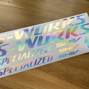 Op de afbeelding: Een witte stickervel met iriserende folielettering. Het vel bevat de woorden "S-Works", "Specialized" en "Shiv" die meerdere keren worden herhaald.
