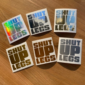 以下が含まれることがあります： 木製の表面に、さまざまな色と仕上げで「Shut Up Legs」というテキストが書かれた6つのステッカーがあります。