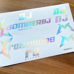 以下が含まれることがあります： 未来的なフォントで「DJ BOMBERDJ marzocchi」の文字が特徴のホログラフィックステッカーシート。ステッカーには、光沢のある虹色の仕上がりで、様式化された「M」と「DJ」のグラフィックも含まれています。