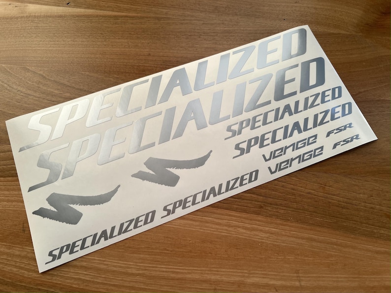 May include: A sheet of silver vinyl decals with the word "Specialized" repeated multiple times in different sizes. The sheet also includes the words "Venge FSR" and a stylized "S" logo.