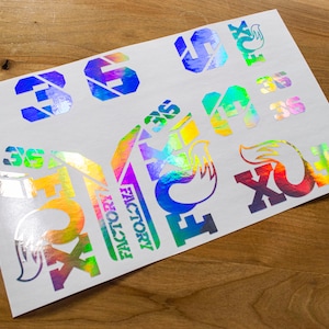 Puede incluir: Una hoja de pegatinas iridiscentes con el logotipo de Fox Racing, las palabras "Factory" y "36", y una silueta de zorro. Las pegatinas son de color arcoíris y tienen un acabado holográfico.