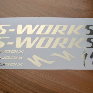 以下が含まれることがあります： 自転車用の金色ビニールステッカーシート。ステッカーには「S-Works」と「Roubaix」の文字と、スタイリッシュな稲妻が描かれています。