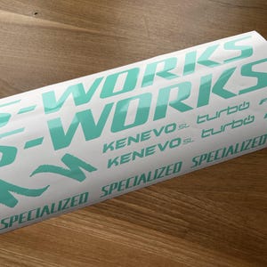 May include: A white sticker with the text "S-WORKS" in teal green lettering. The sticker also includes the text "KENEVOSL turbo FSR" and "SPECIALIZED" in teal green lettering.