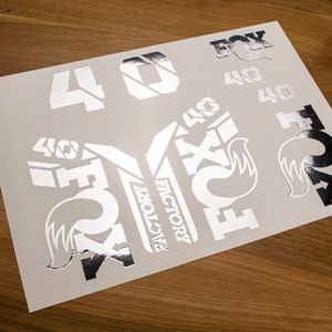 Könnte beinhalten: Eine Blatt mit silbernen Aufklebern mit den Worten "FOX" und "FACTORY" sowie der Zahl "40" in verschiedenen Stilen.