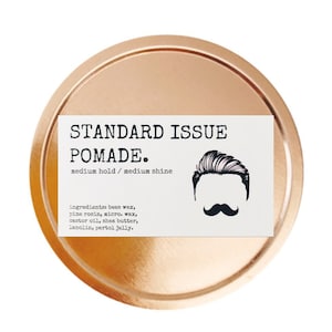Puede incluir: Un recipiente redondo de color dorado con una etiqueta blanca que dice "STANDARD ISSUE POMADE. medium hold / medium shine" y enumera los ingredientes: cera de abejas, resina de pino, micro. cera, aceite de ricino, manteca de karité, lanolina, jalea de petróleo. Una ilustración en blanco y negro de la cara de un hombre con bigote está en la etiqueta.