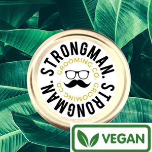May include: A jar with a gold lid featuring the text "STRONGMAN GROOMING CO." in a circular pattern around a logo of glasses and a mustache. The jar is labeled "VEGAN" and set against a backdrop of green banana leaves.