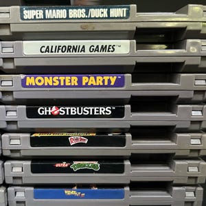 May include: A pile of grey video game cartridges. The labels show game titles including "SUPER MARIO BROS./DUCK HUNT", "CALIFORNIA GAMES", "MONSTER PARTY", and "GHOSTBUSTERS". The cartridges are neatly stacked.