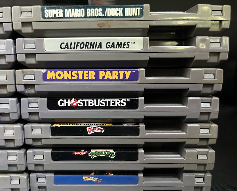 May include: A pile of grey video game cartridges. The labels show game titles including "SUPER MARIO BROS./DUCK HUNT", "CALIFORNIA GAMES", "MONSTER PARTY", and "GHOSTBUSTERS". The cartridges are neatly stacked.