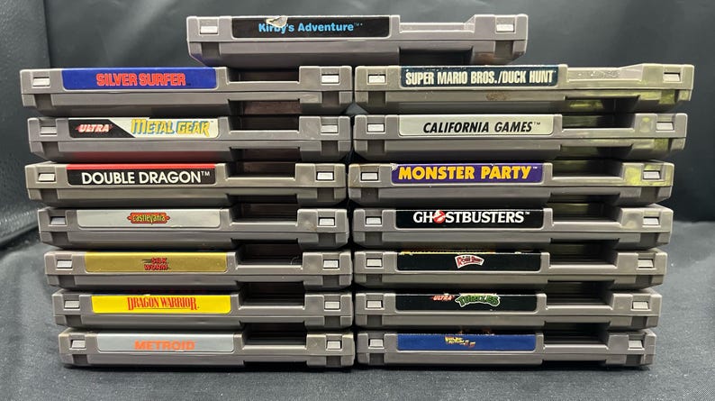 May include: A collection of grey video game cartridges with vibrant labels. Titles include "Kirby's Adventure," "Silver Surfer," "Metal Gear," "Double Dragon," "Dragon Warrior," "Metroid," "Super Mario Bros./Duck Hunt," "California Games," "Monster Party," and "Ghostbusters."