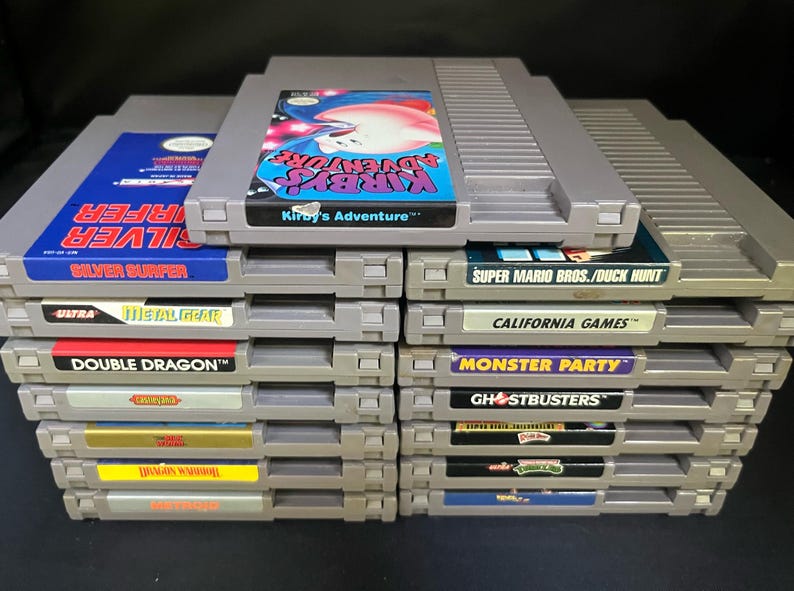 May include: A collection of retro Nintendo Entertainment System (NES) game cartridges. Titles include "Kirby's Adventure," "Silver Surfer," "Double Dragon," and "Super Mario Bros./Duck Hunt." The cartridges are grey with colourful label art.
