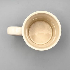 Puede incluir: Taza de cer&aacute;mica color crema con asa redondeada. El interior de la taza es de color beige claro. La taza est&aacute; vac&iacute;a y sobre una superficie gris.
