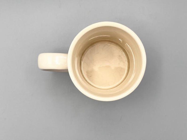 Puede incluir: Taza de cer&aacute;mica color crema con asa redondeada. El interior de la taza es de color beige claro. La taza est&aacute; vac&iacute;a y sobre una superficie gris.
