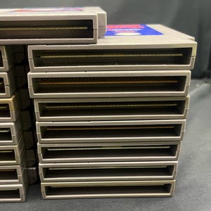 May include: A collection of grey Nintendo Entertainment System (NES) game cartridges. The cartridges are rectangular with a slot at the base and a label on the top. The labels feature blue and red detailing.