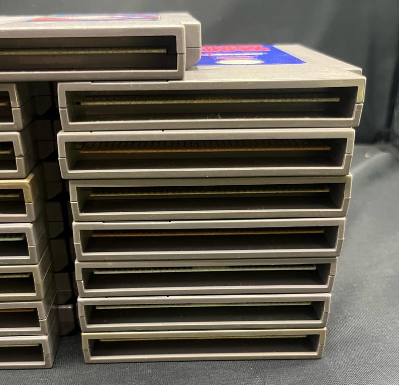May include: A collection of grey Nintendo Entertainment System (NES) game cartridges. The cartridges are rectangular with a slot at the base and a label on the top. The labels feature blue and red detailing.