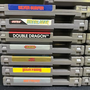 May include: A collection of retro NES game cartridges. Titles visible include "Silver Surfer," "Metal Gear," "Double Dragon," "Castlevania," "Silk Worm," "Dragon Warrior," and "Metroid." The cartridges are grey with colourful labels.