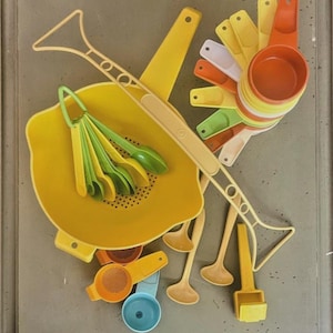 Pode incluir: Uma coleção de utensílios de cozinha coloridos, incluindo xícaras de medição, colheres e um coador. O conjunto apresenta vários tons de amarelo, laranja, verde e branco. Os utensílios são feitos de plástico e estão dispostos em um fundo neutro.