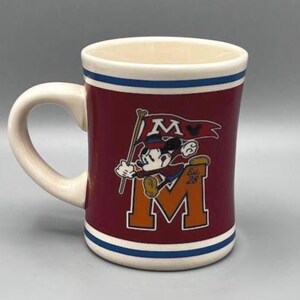 Puede incluir: Una taza de cer&aacute;mica granate con asa y borde blancos, con un dise&ntilde;o de Mickey Mouse sosteniendo una bandera con la letra "M" y el texto "Est. 28". La taza tiene rayas azules y blancas.