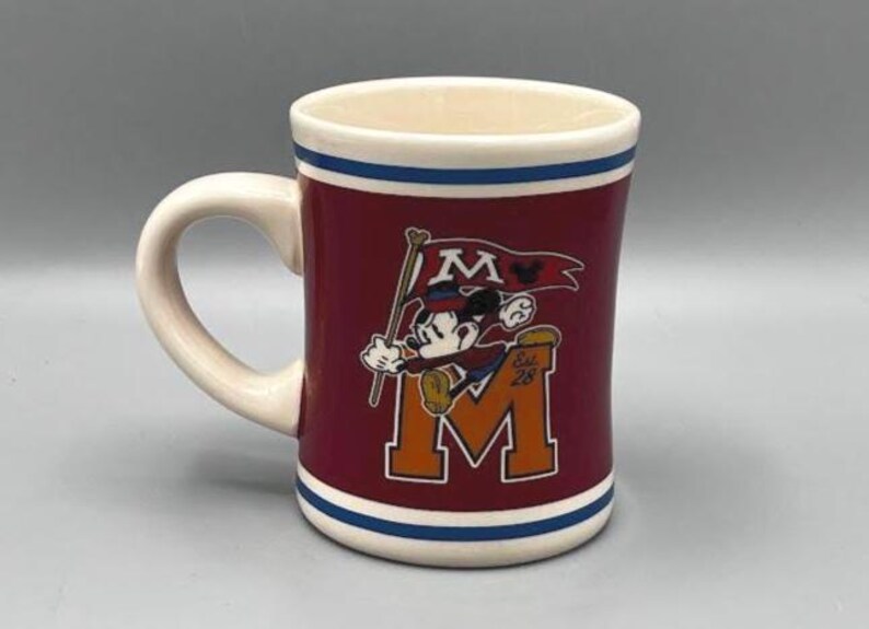 Puede incluir: Una taza de cer&aacute;mica granate con asa y borde blancos, con un dise&ntilde;o de Mickey Mouse sosteniendo una bandera con la letra "M" y el texto "Est. 28". La taza tiene rayas azules y blancas.