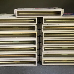 May include: A collection of vintage video game cartridges. The cartridges are rectangular with a light grey casing and gold-coloured connectors. The cartridges are stacked in rows, showcasing a retro gaming theme.