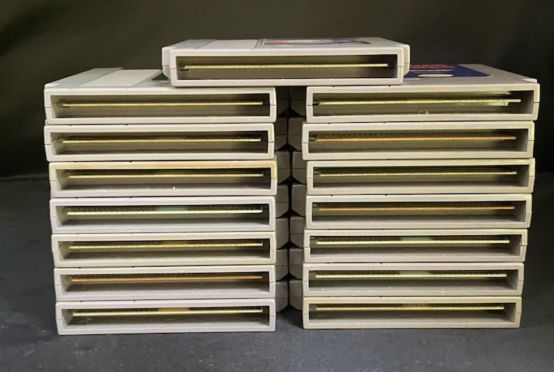 May include: A collection of vintage video game cartridges. The cartridges are rectangular with a light grey casing and gold-coloured connectors. The cartridges are stacked in rows, showcasing a retro gaming theme.