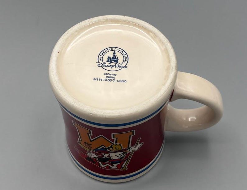 Puede incluir: Una taza de cer&aacute;mica con interior y asa color crema. El exterior presenta una banda granate con una "W" blanca y un personaje de dibujos animados. Rayas azules y blancas acent&uacute;an la parte superior e inferior. La parte inferior de la taza tiene el logotipo de Disney Parks.