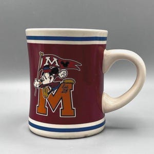 Puede incluir: Taza de cer&aacute;mica granate con asa y borde blancos, con Mickey Mouse sosteniendo una bandera con la letra "M". La taza tiene una franja azul en la base y una gran "M" naranja con "Est. 28".