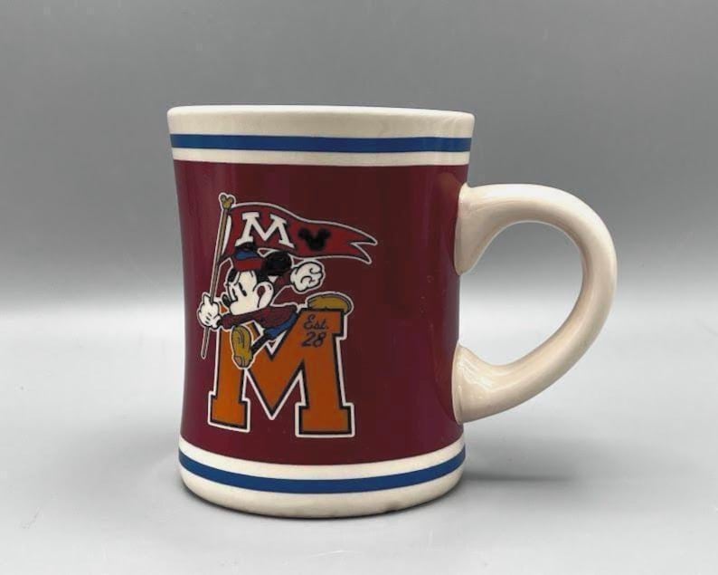 Puede incluir: Taza de cer&aacute;mica granate con asa y borde blancos, con Mickey Mouse sosteniendo una bandera con la letra "M". La taza tiene una franja azul en la base y una gran "M" naranja con "Est. 28".