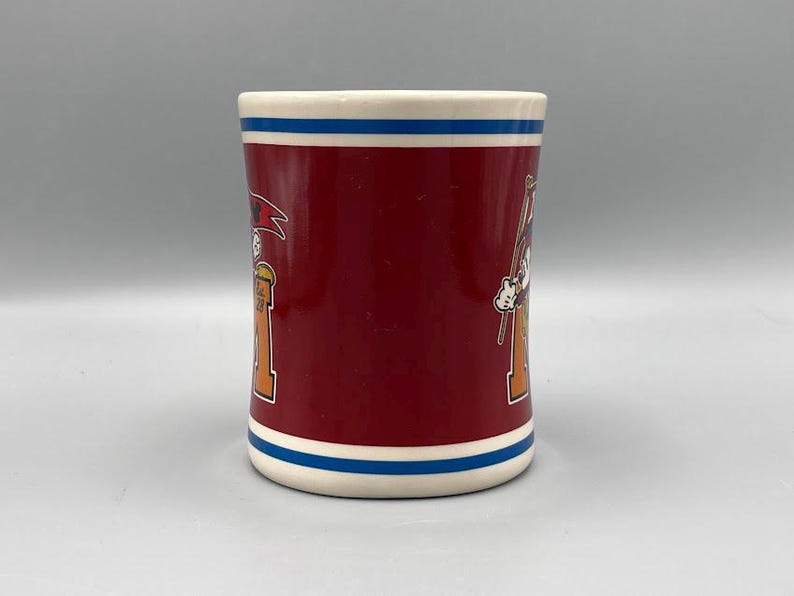 Puede incluir: Una taza de cer&aacute;mica con cuerpo granate, borde blanco y una franja azul cerca de la parte superior e inferior. La taza presenta ilustraciones de dibujos animados de un personaje con sombrero e instrumento musical.