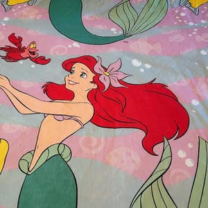 May include: A bed sheet featuring a colorful illustration of Ariel, the Little Mermaid, with her red hair and a flower in her hair. The design includes Flounder, Sebastian, and other aquatic elements on a pink and blue striped background.