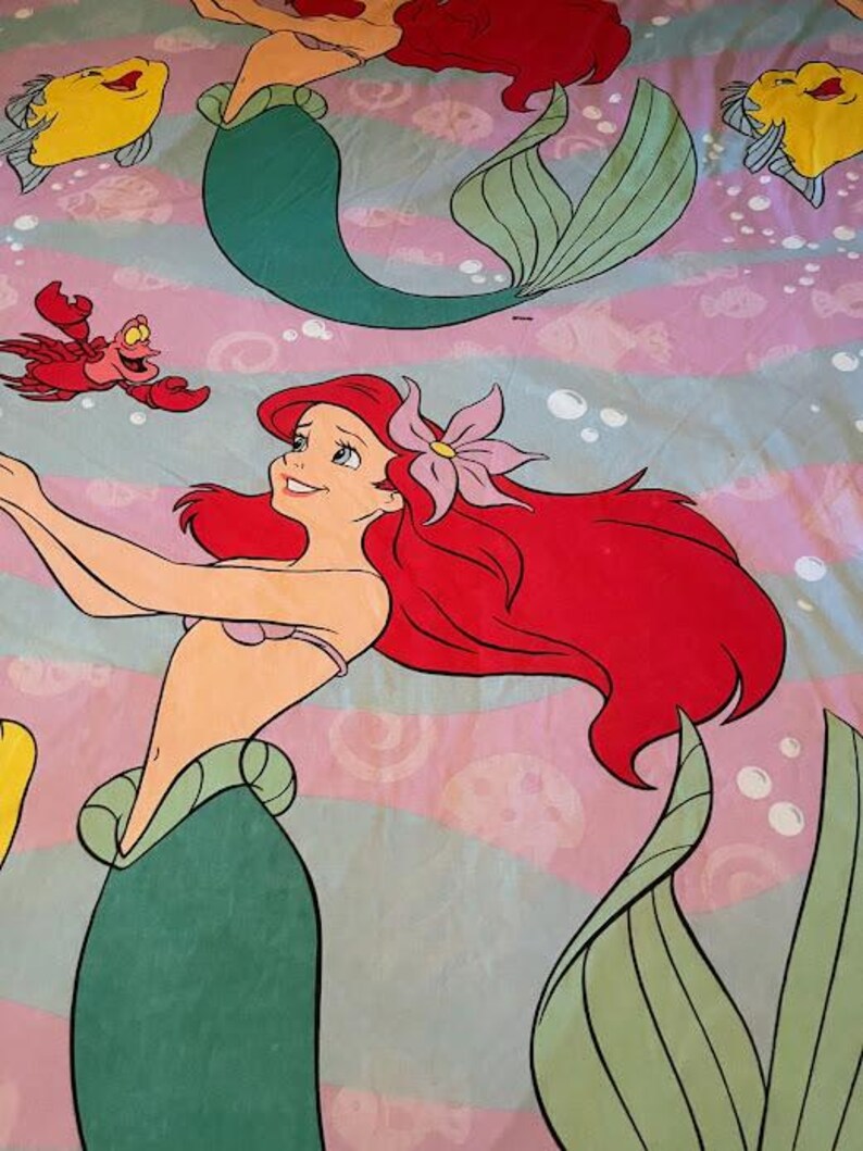 May include: A bed sheet featuring a colorful illustration of Ariel, the Little Mermaid, with her red hair and a flower in her hair. The design includes Flounder, Sebastian, and other aquatic elements on a pink and blue striped background.
