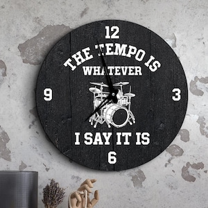 May include: A round, black wooden wall clock with white text and a drum set graphic. The text reads "The Tempo Is Whatever I Say It Is." Numbers 12, 3, 6, and 9 are displayed. The clock has a distressed wood grain appearance.