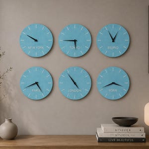 May include: A set of six circular wall clocks with light blue faces, white hour markers, and black hands. Each clock features a city name printed in white text: New York, Tokyo, Beijing, Paris, London, and Rome. The clocks are arranged in two rows.