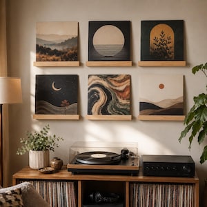 May include: Six square art prints displayed on two wooden wall shelves. The prints feature various landscape and abstract designs including a mountain range, a large sun, a botanical arch, a crescent moon, swirling colours, and a desert scene.