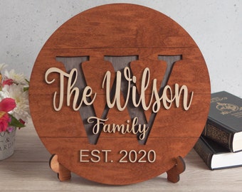 Könnte beinhalten: Ein rundes Holzschild mit einem großen ausgeschnittenen "W". Das Schild ist braun lackiert und trägt den Text "The Wilson Family EST. 2020" in hellbrauner Farbe.
