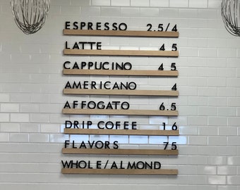 May include: A wall-mounted menu board featuring horizontal light wood slats with black lettering. The text lists coffee items including Espresso, Latte, Cappuccino, Americano, Affogato, Drip Coffee, Flavors, and Whole/Almond with corresponding prices.