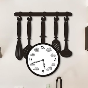 Peut inclure: Une horloge murale noire avec un cadran circulaire affichant des chiffres noirs et de petites illustrations de nourriture. Au-dessus du cadran, un support métallique noir soutient cinq ustensiles de cuisine suspendus, dont un fouet et une louche.