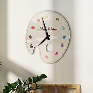 May include: A wall clock shaped like a painter's palette, with colorful paint splotches around the edge. The clock face is off-white, with black hands and the name "Mrs. Watson" printed on it. The clock is mounted on a white wall.
