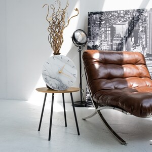 May include: A white marble clock with black markings and gold hands sits on a small wooden side table with black metal legs. A brown leather chaise lounge with a silver frame is in the background.