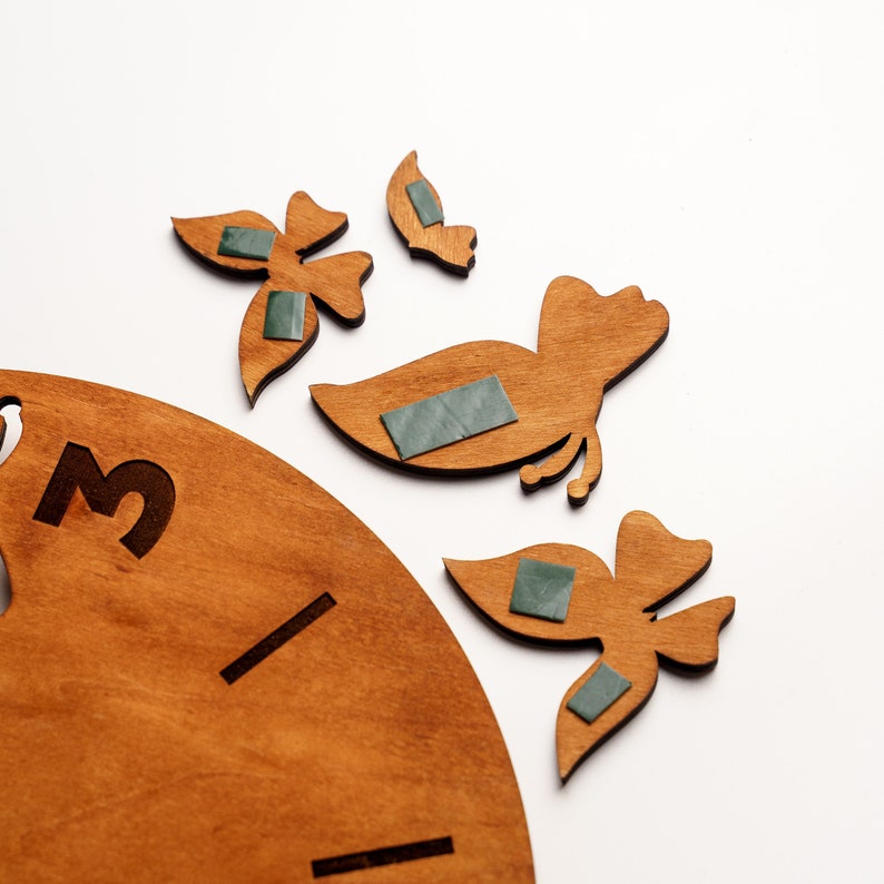 May include: A wooden clock face with the number 3 and a black line. The clock is surrounded by four wooden butterfly shapes with green adhesive backing.