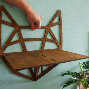 May include: A wooden wall shelf with a geometric design in the shape of a fox head. The shelf is brown and has a light brown stain. The shelf is mounted on a light blue wall.