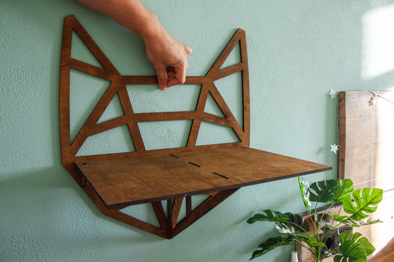 May include: A wooden wall shelf with a geometric design in the shape of a fox head. The shelf is brown and has a light brown stain. The shelf is mounted on a light blue wall.