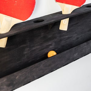 May include: A black wooden wall-mounted rack with two red and black ping pong paddles and a single orange ping pong ball.