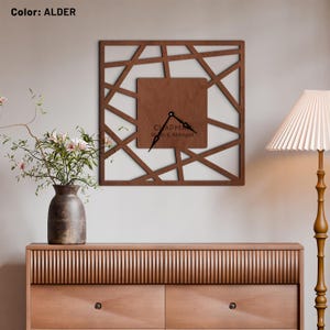 Puede incluir: Un reloj de pared cuadrado de madera con un diseño geométrico. La esfera del reloj es de un tono marrón más claro, con manecillas negras y el texto "CHAPMAN Ryan & Abigail". El reloj está montado en una pared gris claro.