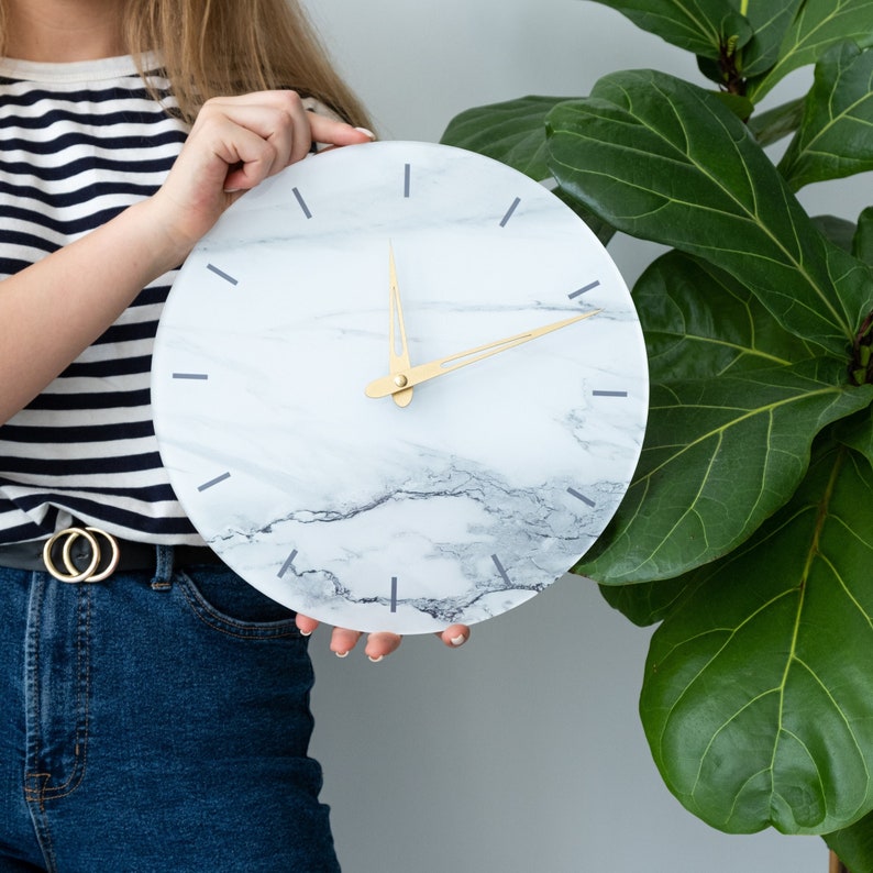 May include: A white marble clock with gold hands and black markings. The clock is round and has a white background with gray marble veins.