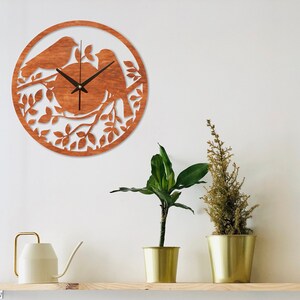 May include: A wooden wall clock with a brown finish features a silhouette of two birds perched on a branch with leaves. The clock face is visible with black hands and a white background.