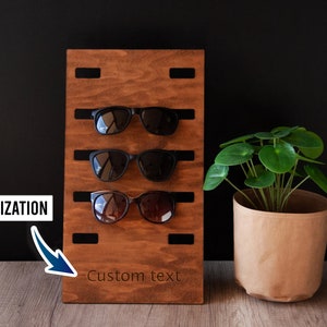 Puede incluir: Un soporte de madera marr&oacute;n oscuro para gafas de sol con tres pares de gafas de sol. El soporte tiene ranuras para sujetar las gafas. El texto "Personalizaci&oacute;n" y "Custom text" son visibles.